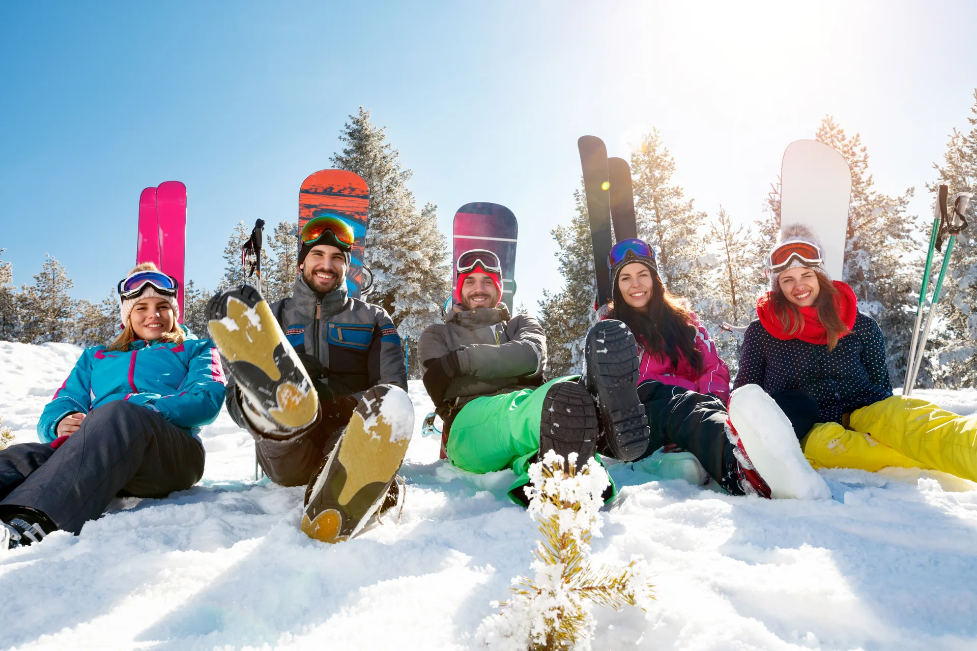 Five friends laughing in the snow with skis and snowboards held upright