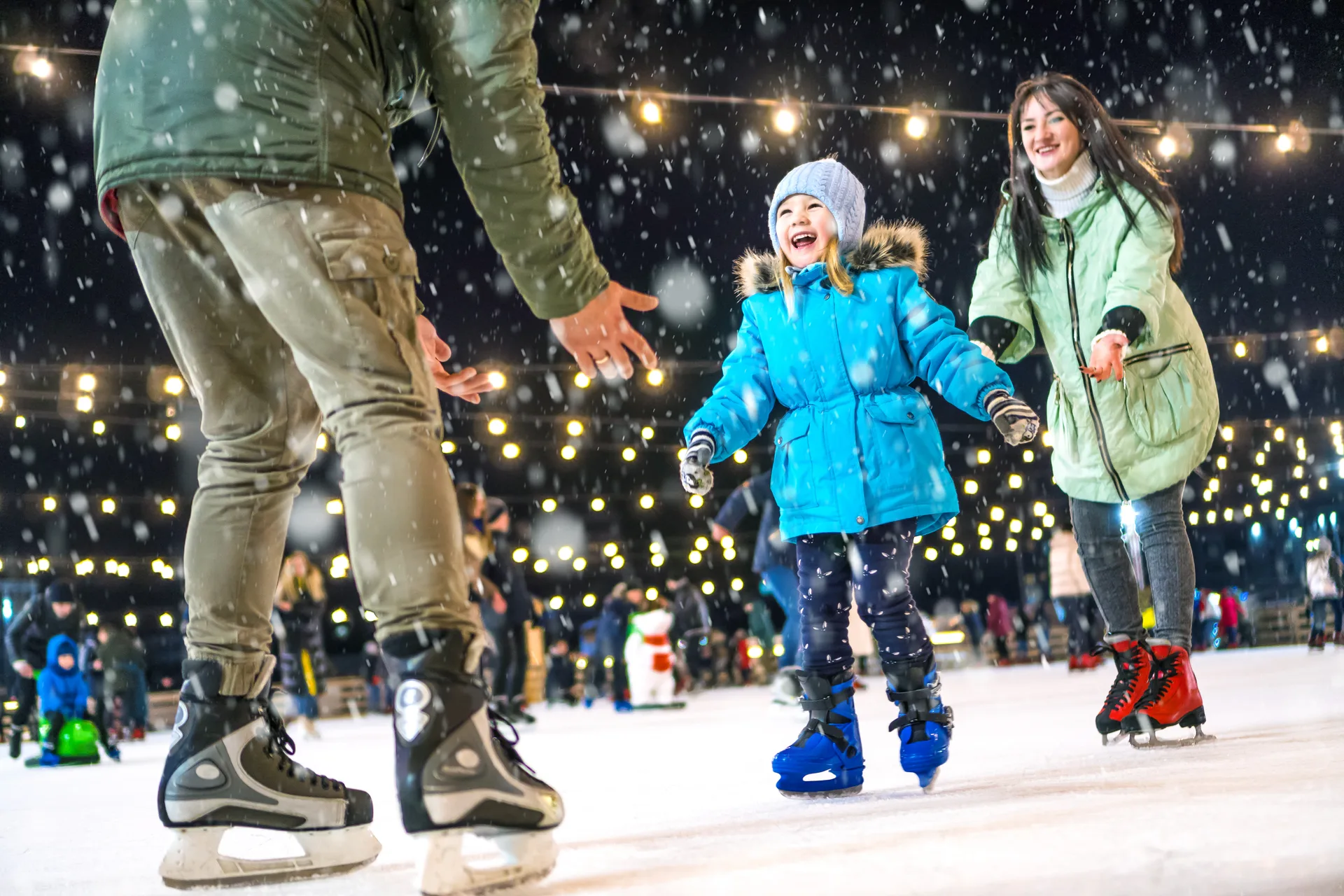 Parents helping a young girl skate on a string-lit outdoor rink in snowfall