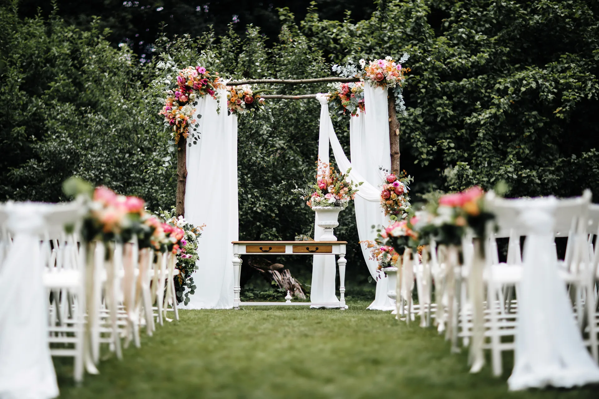 Outdoor wedding ceremony aisle with white chairs, a flower-decorated wooden arch and white drapes