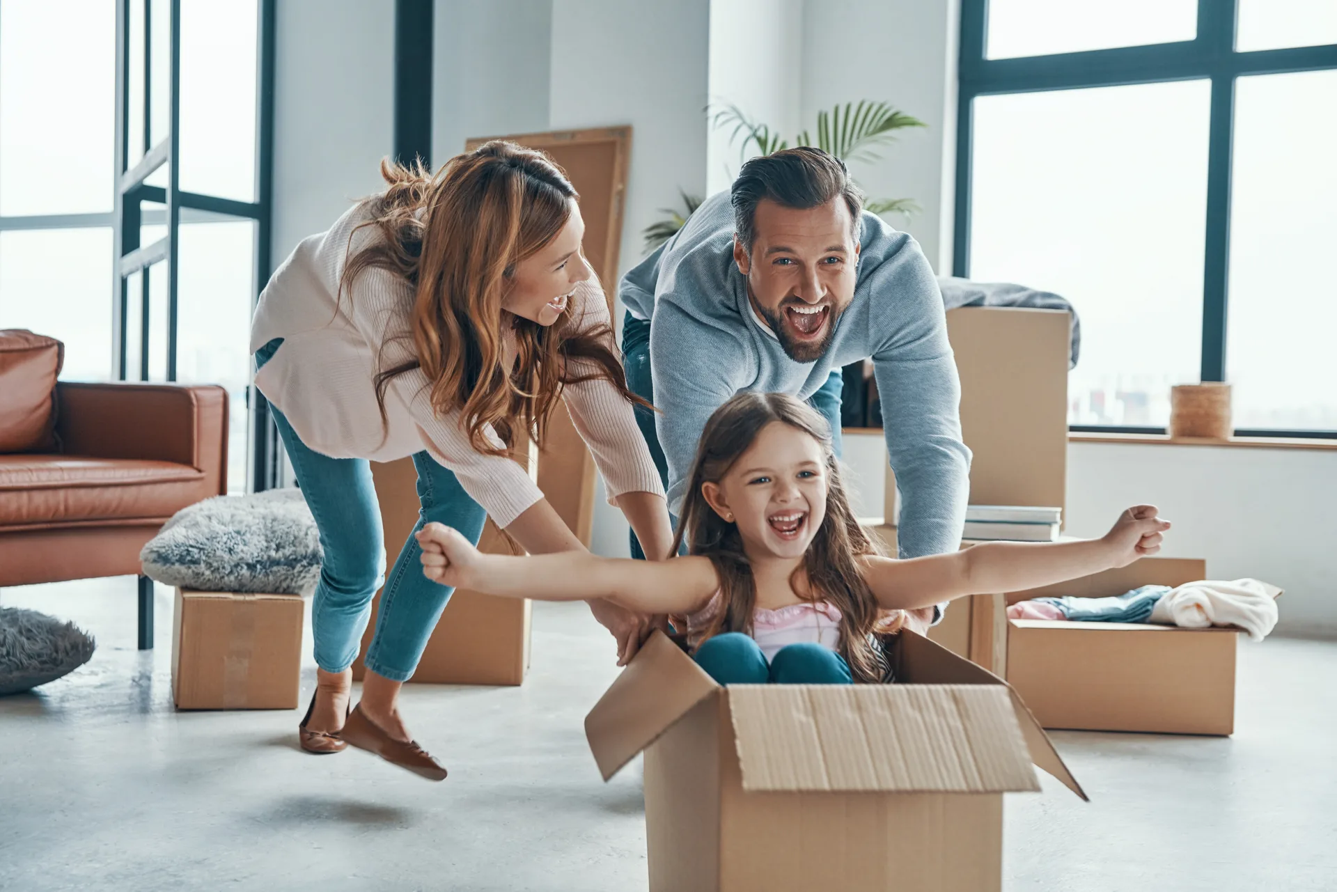 Parents laughing as they push their young daughter across the floor in a cardboard box