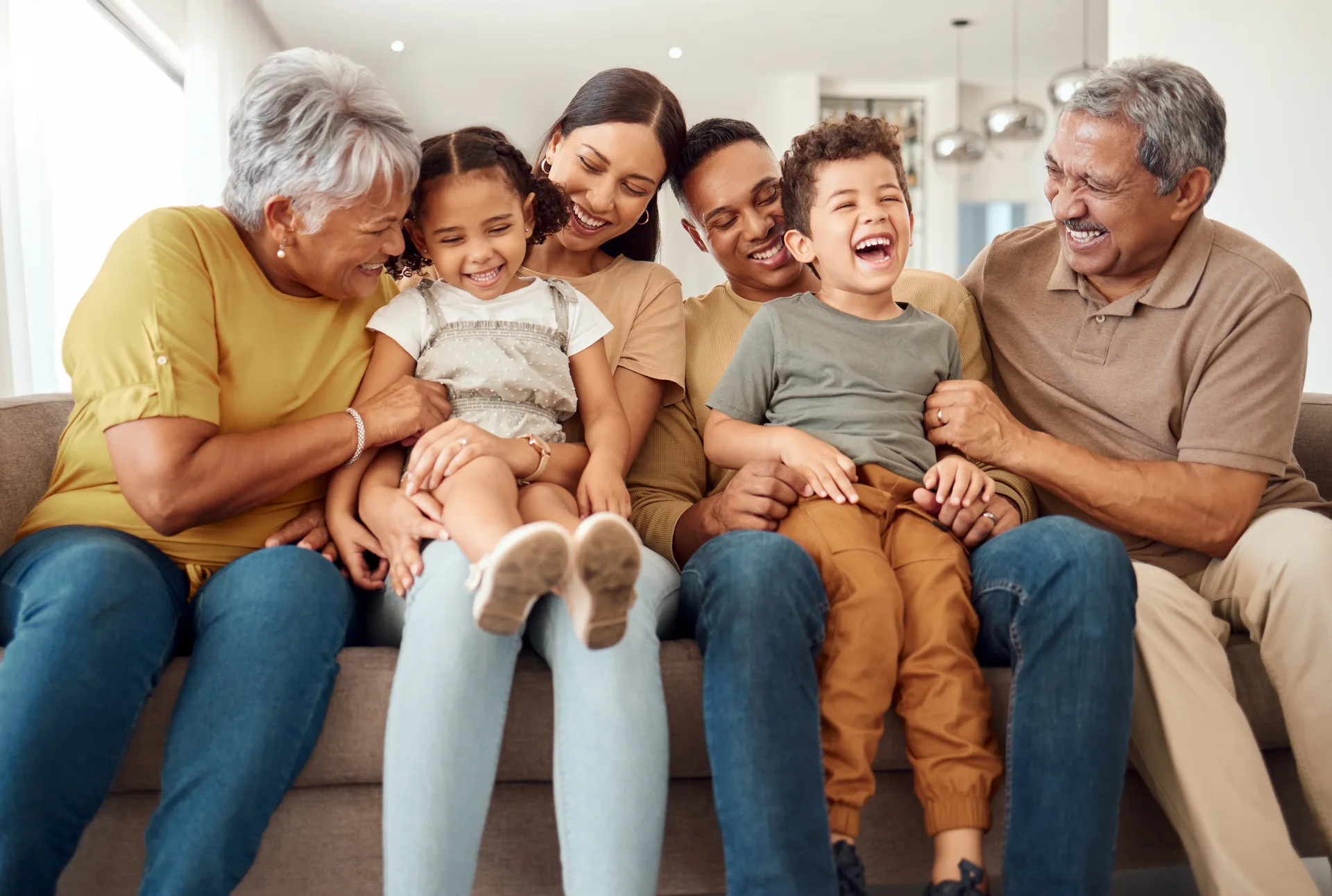 Grandparents, parents and two laughing children sitting close together on a beige sofa