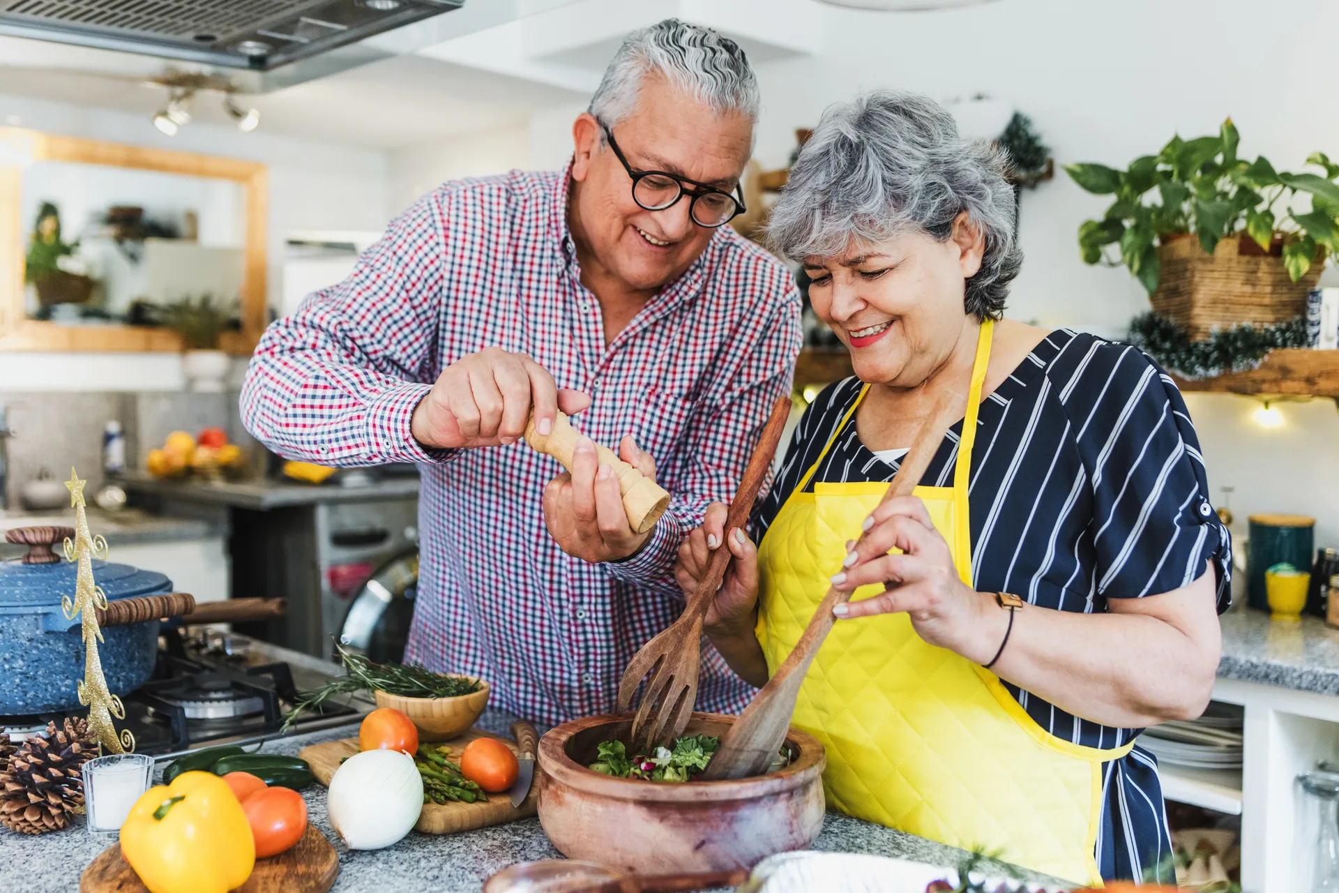 Older couple in a home kitchen tossing a salad in a wooden bowl beside fresh vegetables