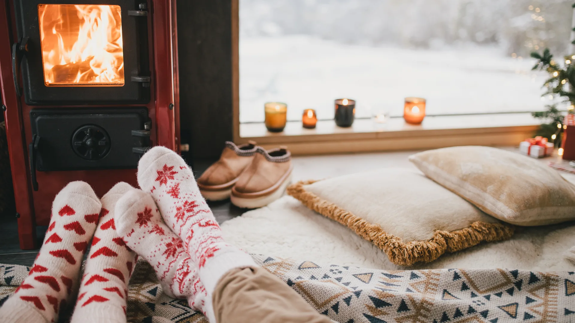 Two pairs of feet in knit socks warming by a wood stove with snow outside