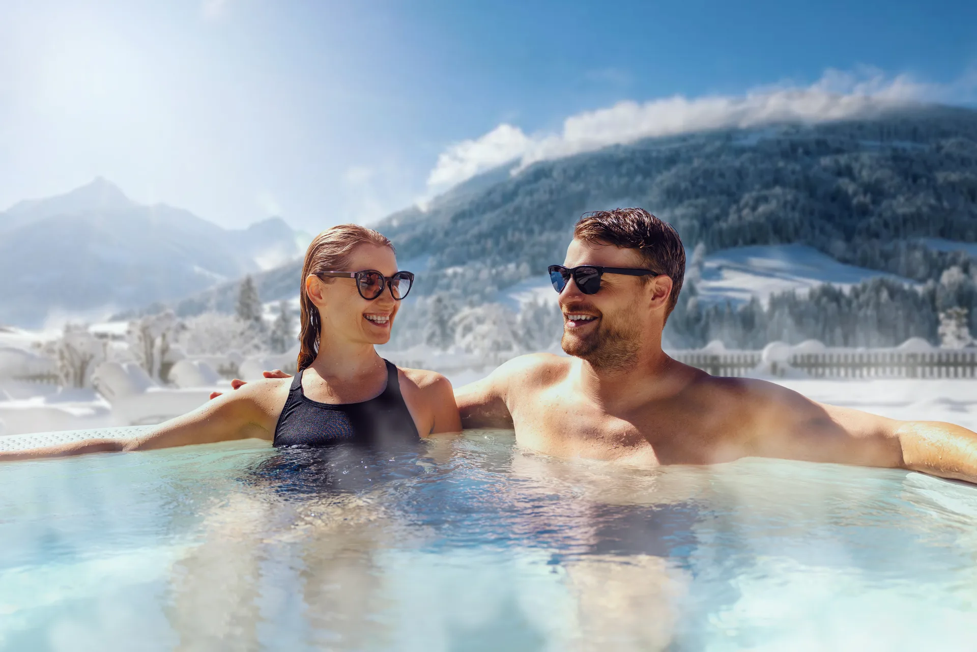 Couple in sunglasses relaxing in a steaming outdoor hot tub with snowy mountains behind