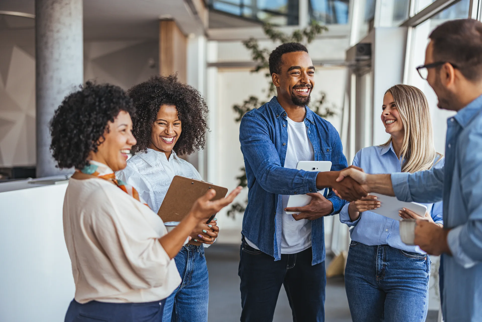 Five colleagues in jeans and shirts shaking hands and smiling in a bright office lobby