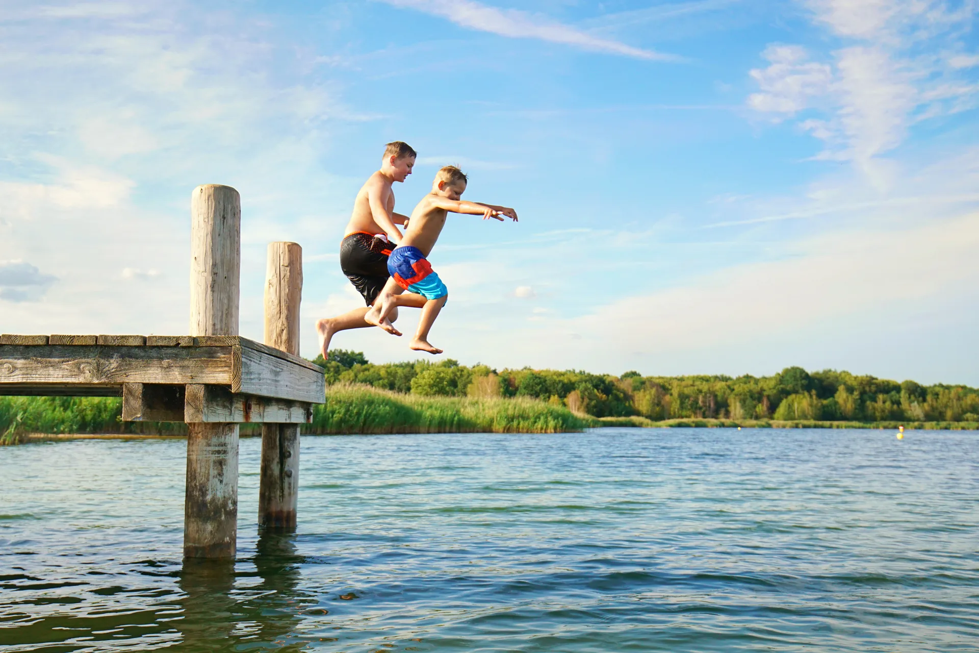Two kids leaping from a wooden jetty into a lake on a summer day