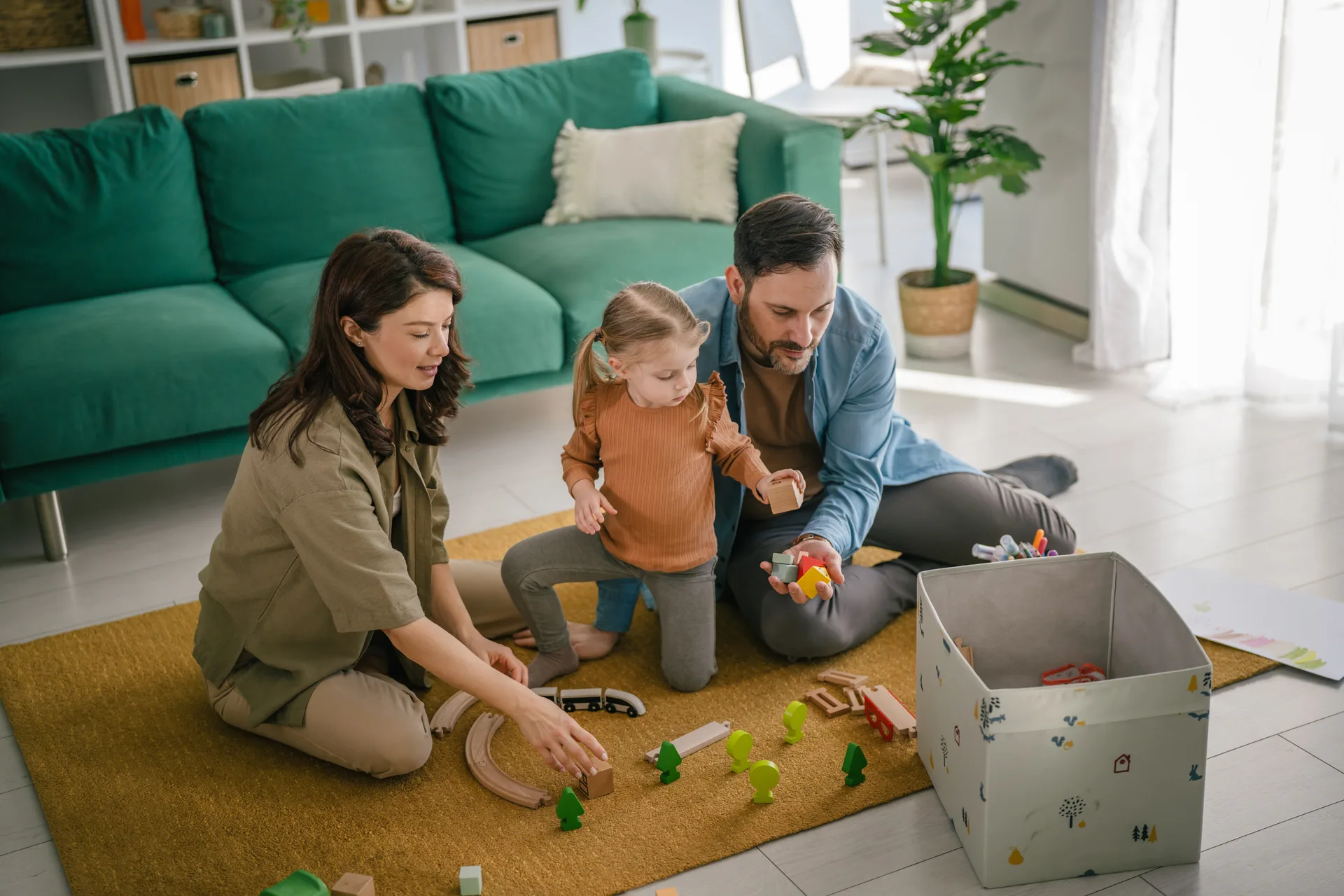 Parents and a toddler playing with a wooden train set on a rug in front of a green sofa