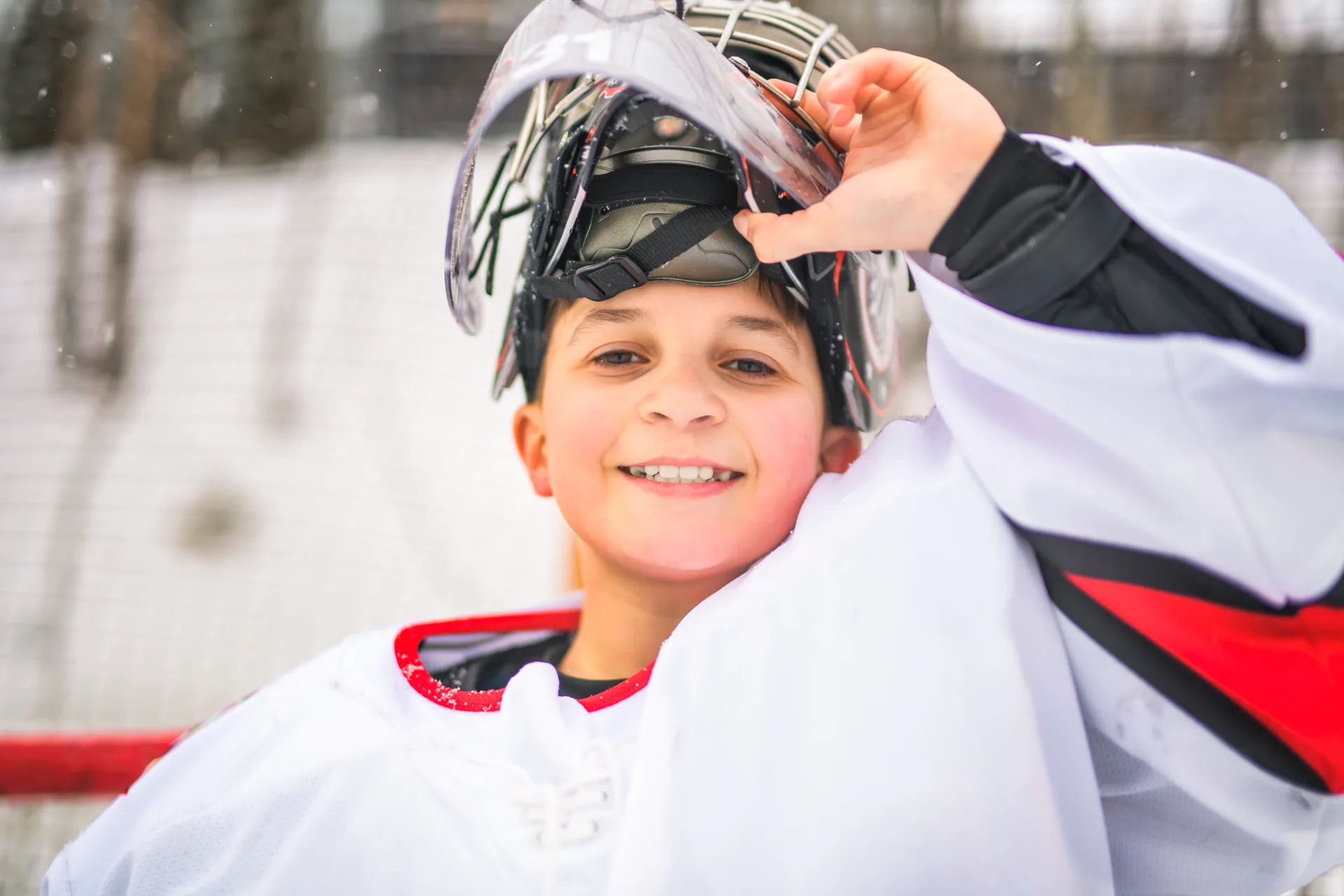 Young goalie in a white hockey jersey lifting his mask and smiling on an outdoor rink