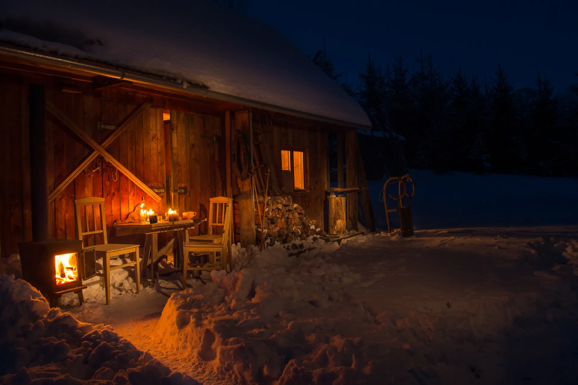 Candle-lit table and wood stove outside a timber cottage on a winter night