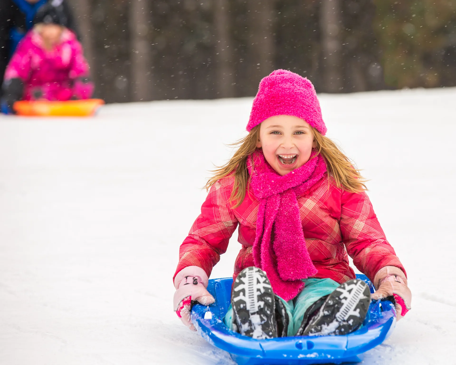 Laughing girl in pink winter gear riding a blue sled downhill