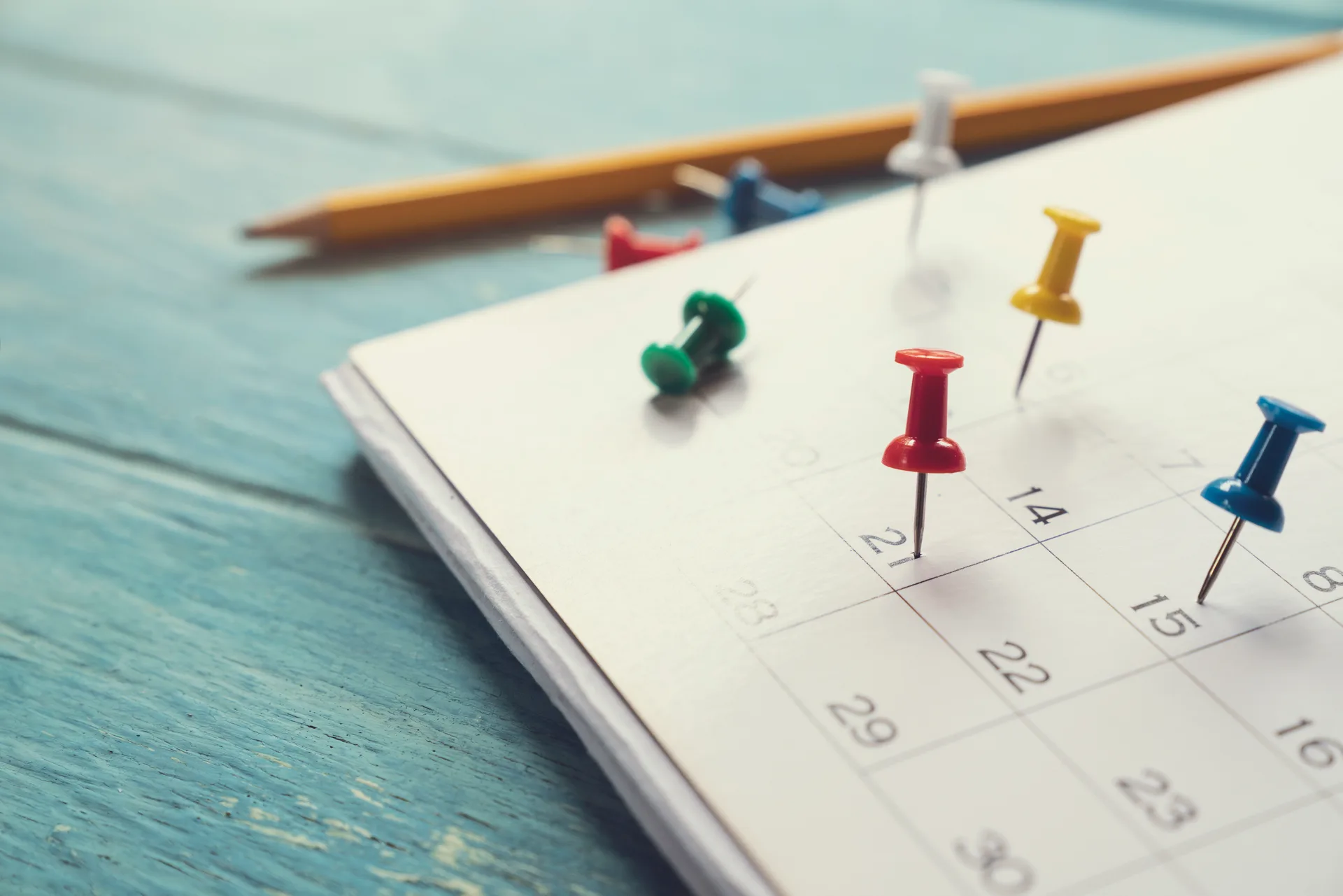 Coloured push pins and a pencil on a paper calendar page lying on a blue wooden table